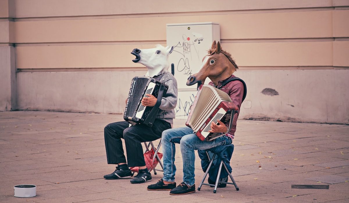 two person wearing horse heads sitting on folding chairs while playing accordions beside brown concrete building